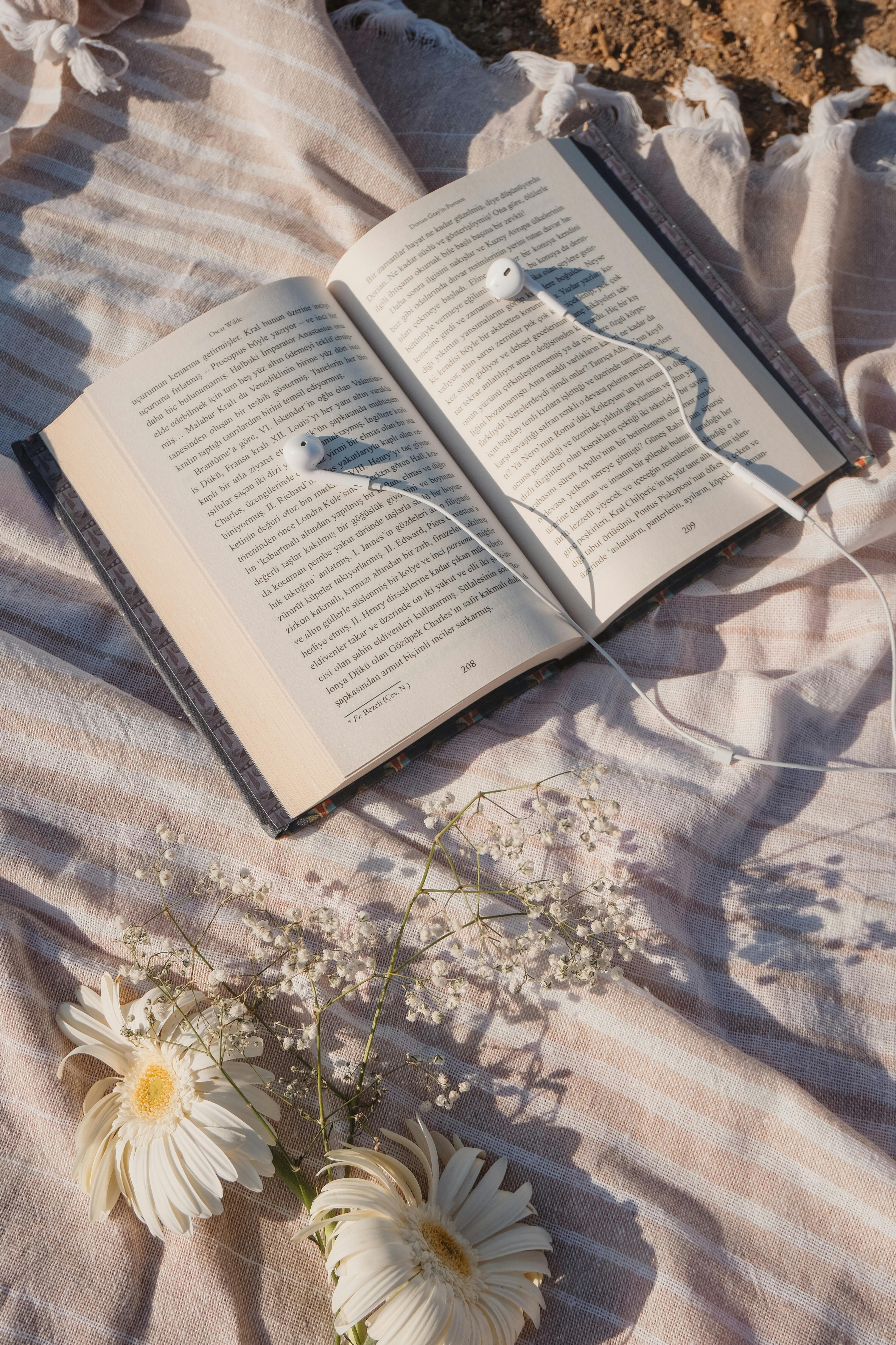 An open book with earphones on a striped blanket, accented by white flowers, in sunlight.